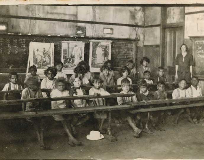 一张旧的黑白照片，教室里的学生和老师坐在课桌前