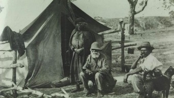 两名土著男子和一名土著妇女站在帐篷前