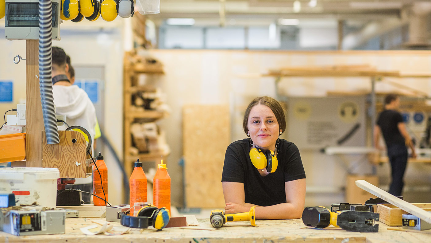肖像微笑年轻女木工学员靠在工作台与电动工具在照明车间-库存照片
